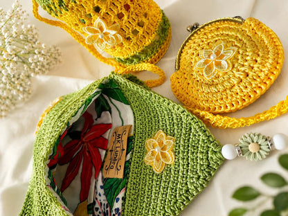 A set of handmade crocheted accessories, including a green textured shoulder bag with a floral applique, a round yellow clutch with a metal clasp, and a yellow-and-green crocheted pouch, arranged on a white fabric background with dried flowers and green leaves.